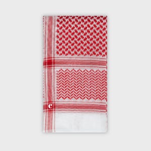 Puede incluir: Un pañuelo keffiyeh blanco y rojo con un patrón geométrico. El pañuelo presenta un patrón repetitivo de cuadrados rojos y líneas diagonales, con un borde rojo. El pañuelo está doblado y se muestra sobre un fondo gris claro.