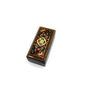 May include: A small, rectangular box with a black base and a colorful, floral design on the lid. The design features intricate details and a variety of colors, including red, yellow, blue, and green.