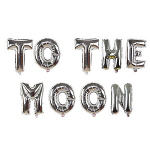 May include: Silver foil balloons spelling out "TO THE MOON". The balloons are inflated and arranged in two rows. The letters are capital and have a metallic, reflective surface, ideal for a party or celebration.