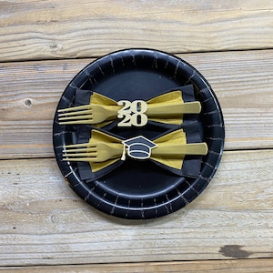 May include: Two gold forks with black bow ties and gold 2020 and graduation cap accents on a black paper plate.