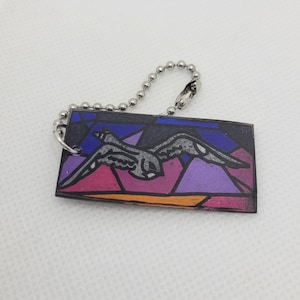 May include: A rectangular keychain with a stained-glass style design featuring a bird in flight. The artwork uses shades of purple, pink, orange, and blue. The keychain has a silver ball chain attached.