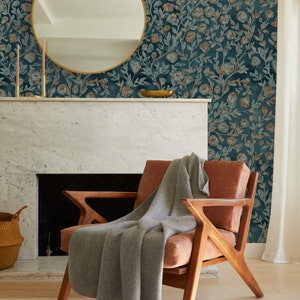 May include: A brown leather armchair with a grey knitted throw blanket. The chair is in front of a fireplace with a white marble mantel and a round gold mirror hanging above it. The wall behind the chair is covered in teal wallpaper with a floral pattern.