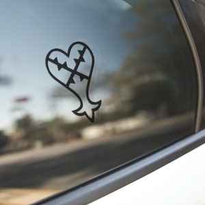 May include: Black heart-shaped decal with a stylized design, affixed to a car window. The decal features a heart shape with a jagged top and a fish-like tail. The design is a solid black color.