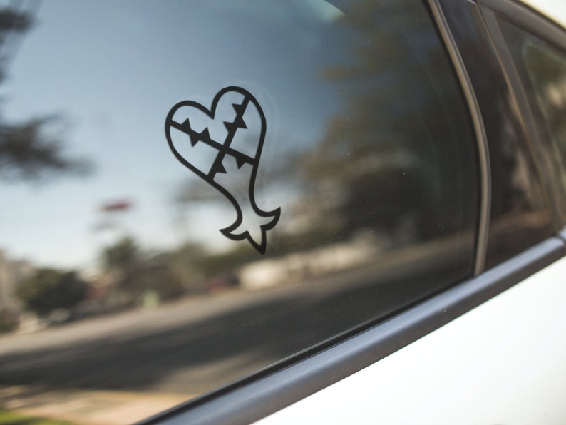May include: Black heart-shaped decal with a stylized design, affixed to a car window. The decal features a heart shape with a jagged top and a fish-like tail. The design is a solid black color.