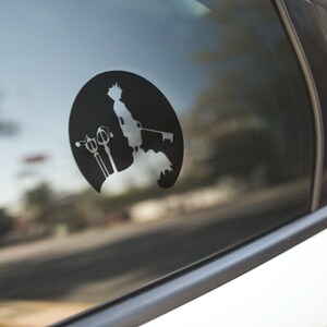 May include: Black vinyl decal featuring a silhouette of a character holding a keyblade, set against a circular background. The design is affixed to a car window, showcasing a detailed illustration of a video game character.