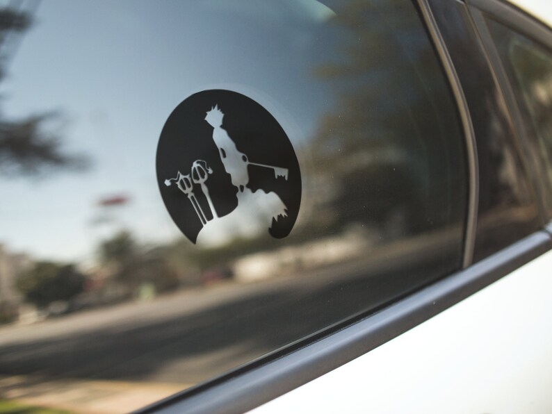 May include: Black vinyl decal featuring a silhouette of a character holding a keyblade, set against a circular background. The design is affixed to a car window, showcasing a detailed illustration of a video game character.
