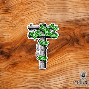 May include: A sticker featuring a stylized illustration of a utility pole with green dollar bills in place of leaves. The text "MONEY TREE" is printed vertically on the pole. The sticker is on a wooden surface.