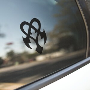 May include: A black vinyl decal featuring an abstract design, affixed to a car window. The design is a stylized combination of hearts and arrowheads. The car window reflects the outside environment, including trees and a street.