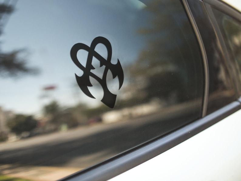 May include: A black vinyl decal featuring an abstract design, affixed to a car window. The design is a stylized combination of hearts and arrowheads. The car window reflects the outside environment, including trees and a street.