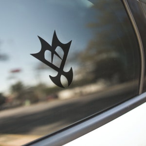 May include: Black vinyl car decal featuring a stylized emblem. The design is a combination of geometric shapes, including a crown-like top and a teardrop-shaped bottom. The decal is affixed to a car window, with the background showing a blurred street scene.