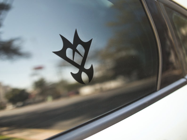 May include: Black vinyl car decal featuring a stylized emblem. The design is a combination of geometric shapes, including a crown-like top and a teardrop-shaped bottom. The decal is affixed to a car window, with the background showing a blurred street scene.
