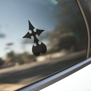 May include: A black vinyl decal of a stylized emblem, affixed to the window of a car. The emblem features a four-pointed star shape with three circular cutouts and a heart-shaped base. The car is white.