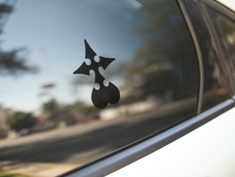 May include: A black vinyl decal of a stylized emblem, affixed to the window of a car. The emblem features a four-pointed star shape with three circular cutouts and a heart-shaped base. The car is white.