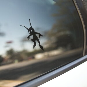 May include: A black vinyl decal of a stylized creature with a rounded body, two antennae, and two legs is affixed to a car window. The creature has a simple, cartoonish design. The background shows a blurred street scene.