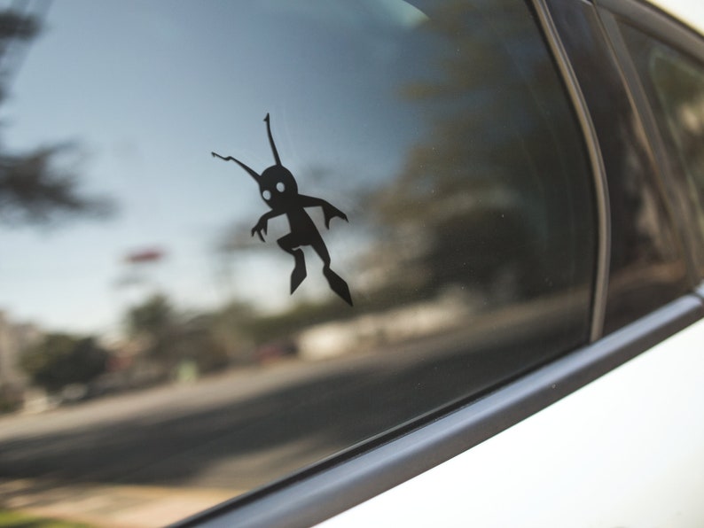May include: A black vinyl decal of a stylized creature with a rounded body, two antennae, and two legs is affixed to a car window. The creature has a simple, cartoonish design. The background shows a blurred street scene.