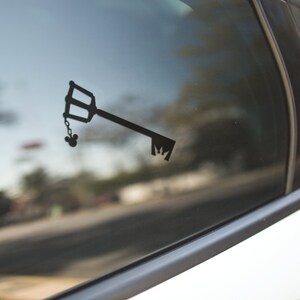May include: Black vinyl decal of a key with a Mickey Mouse head charm, affixed to a car window. The key has a distinctive handle and teeth design. The decal is a silhouette, creating a minimalist aesthetic.