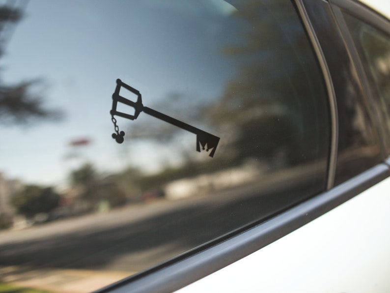 May include: Black vinyl decal of a key with a Mickey Mouse head charm, affixed to a car window. The key has a distinctive handle and teeth design. The decal is a silhouette, creating a minimalist aesthetic.