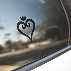 May include: Black heart-shaped decal with a crown on top, affixed to a car window. The design features a stylized heart with decorative swirls and a small crown. The decal is a solid black color and is visible against the reflective glass.