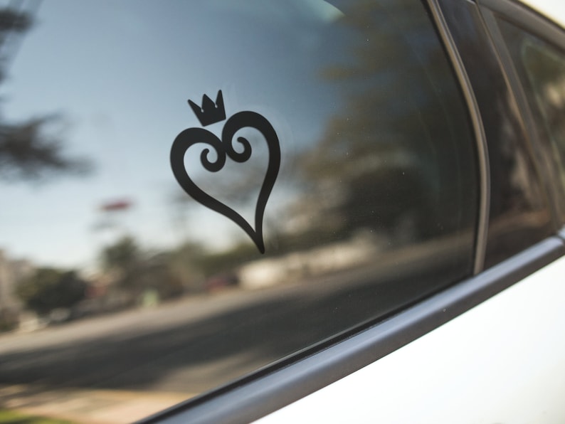 May include: Black heart-shaped decal with a crown on top, affixed to a car window. The design features a stylized heart with decorative swirls and a small crown. The decal is a solid black color and is visible against the reflective glass.