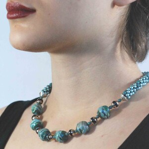 May include: A teal and silver beaded necklace. The necklace features large, marbled teal beads interspersed with smaller silver and dark beads. The necklace is worn against a black top.