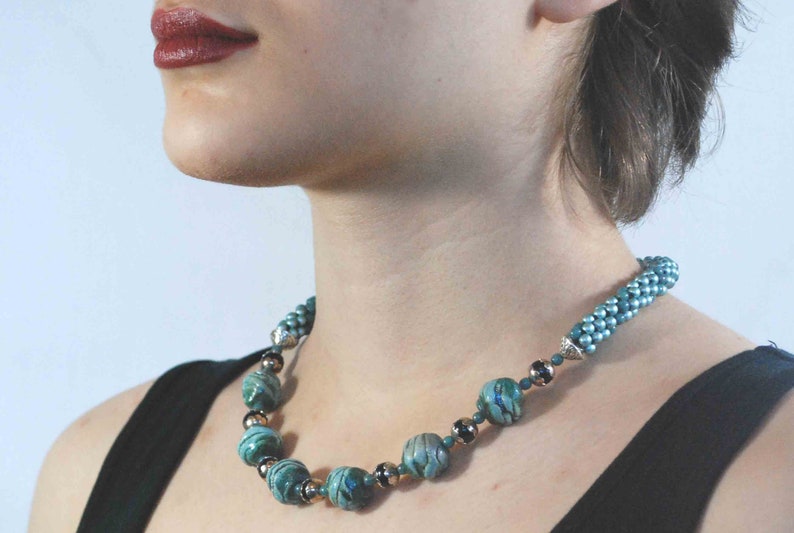 May include: A teal and silver beaded necklace. The necklace features large, marbled teal beads interspersed with smaller silver and dark beads. The necklace is worn against a black top.