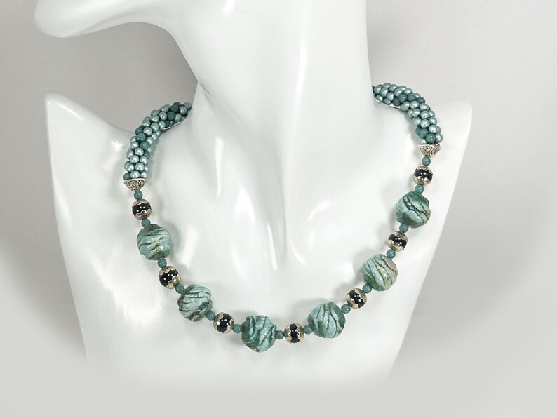 May include: A teal beaded necklace on a white mannequin bust. The necklace has large, marbled beads in teal and green, with smaller black and silver beads. The upper part of the necklace is a tightly woven strand of small teal beads.