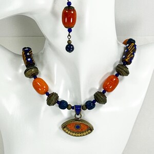 May include: A necklace and earring set featuring orange and blue beads, brass accents, and an eye-shaped pendant. The necklace and earrings are displayed on a white mannequin bust. The earrings have a single orange bead and blue accents.
