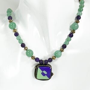 May include: A beaded necklace featuring green and purple beads with gold-colored spacers. The pendant is square, with a green and purple geometric design. The necklace is displayed on a white mannequin.