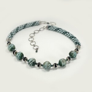 May include: A beaded necklace with a mix of green and silver beads. The necklace has a braided design with larger, marbled green beads interspersed. The clasp is silver-toned, and the necklace is displayed on a white background.