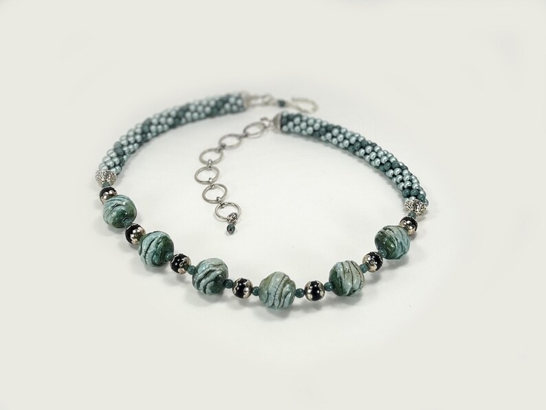 May include: A beaded necklace with a mix of green and silver beads. The necklace has a braided design with larger, marbled green beads interspersed. The clasp is silver-toned, and the necklace is displayed on a white background.