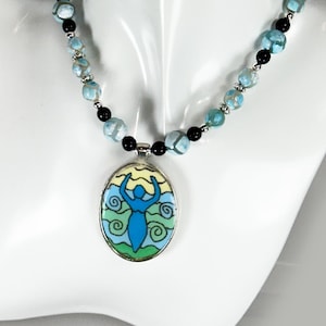 May include: A necklace featuring a central oval pendant with a blue stylized figure against a yellow, green, and blue background. The chain consists of alternating light blue marbled beads, small black beads, and silver-toned metal spacers.