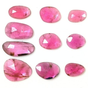 Può includere: Una collezione di otto pietre preziose rosa sfaccettate e sciolte disposte in una griglia. Ogni pietra presenta una forma arrotondata, irregolare e unica con una superficie lucida e riflettente che mostra inclusioni interne e varie tonalità di rosa magenta vibrante.