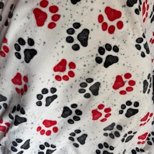 Puede incluir: Manta de forro polar blanca con un estampado de huellas de patas negras y rojas y pequeñas motas grises. Las huellas están dispersas por la superficie, creando un diseño lúdico. La manta es suave.