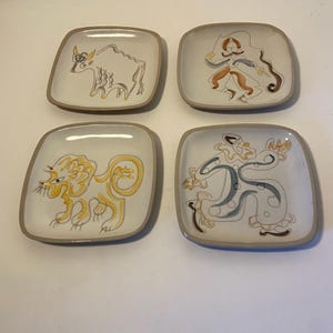 May include: Set of four square ceramic plates with rounded corners. Each plate features a unique hand-drawn illustration in shades of yellow, brown, and blue on a white background, with a brown border. The illustrations depict stylized figures and animals.