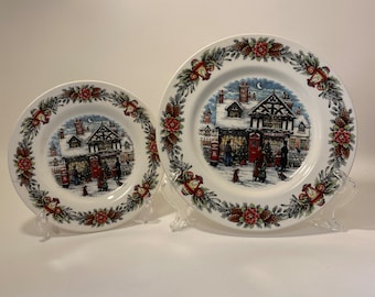 May include: Two white ceramic plates with a winter scene of a building with red doors and snow on the roof. The plates have a green and red border with holly and pine cones.