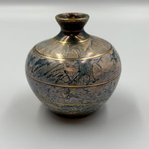 Peut inclure: Petit vase décoratif au corps arrondi et au col étroit. Le vase présente un motif marbré dans les tons de gris, de bleu et d'or. Des bandes dorées encerclent le vase, ajoutant une touche d'élégance. Le dessus du vase est brun foncé.