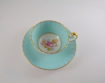 May include: A light blue teacup and saucer set with a gold trim and a floral design. The cup has a white interior with a pink rose and gold accents.