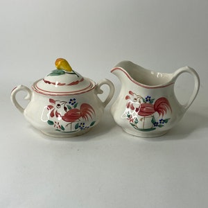 May include: A white ceramic creamer and sugar bowl set with a rooster design. The rooster is painted in shades of pink, red, green, and blue. The creamer has a handle and a spout. The sugar bowl has a lid with a yellow pear finial.