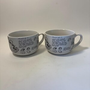 Puede incluir: Dos tazas de cer&aacute;mica blanca con asas. Cada taza presenta un patr&oacute;n de cuadr&iacute;cula y textos y bocetos manuscritos de Minnie Mouse. El texto de las tazas describe la construcci&oacute;n y el disfraz de Minnie.