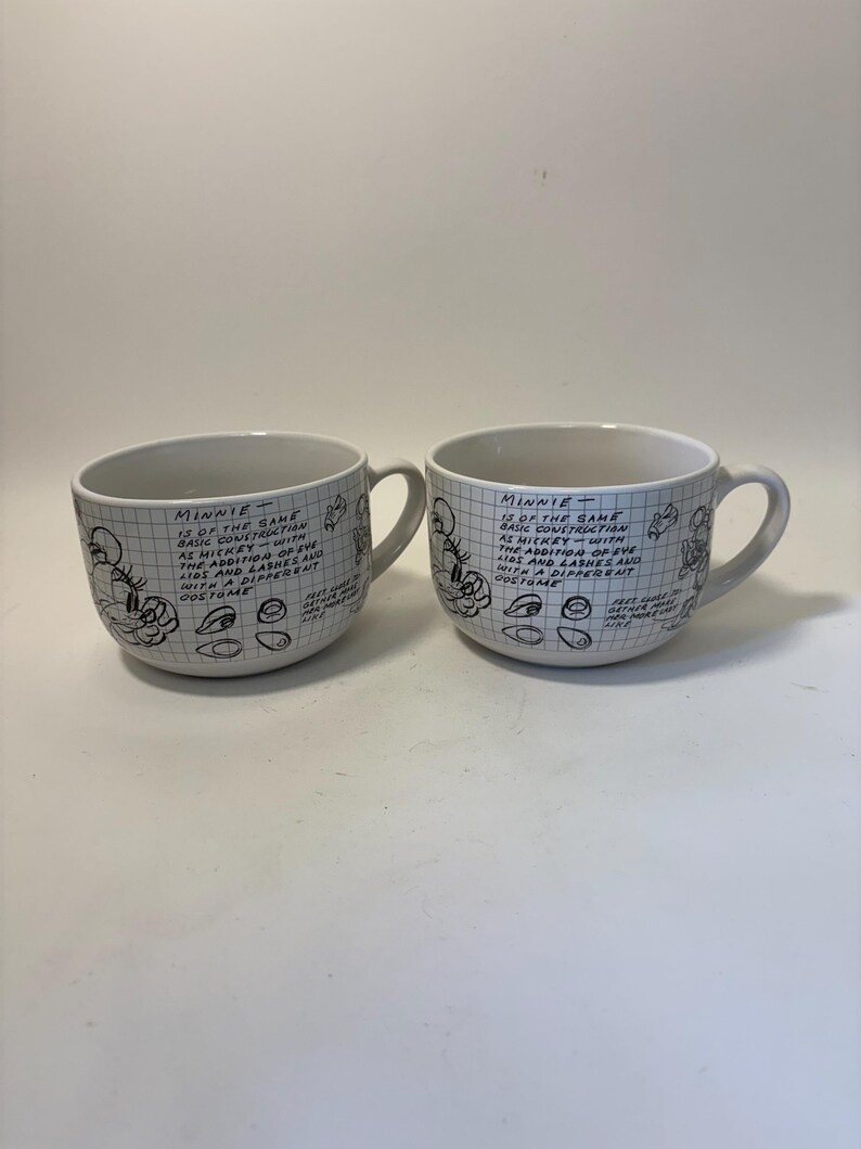 Puede incluir: Dos tazas de cer&aacute;mica blanca con asas. Cada taza presenta un patr&oacute;n de cuadr&iacute;cula y textos y bocetos manuscritos de Minnie Mouse. El texto de las tazas describe la construcci&oacute;n y el disfraz de Minnie.