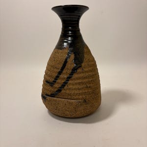 May include: A handmade ceramic vase with a textured, brown body and a black, glossy neck. The vase features a dark, dripped design. The vase is a unique piece of pottery.
