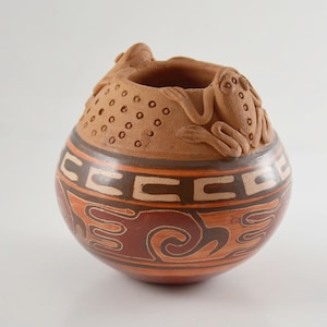 Peut inclure: Un pot en terre cuite brun avec un motif géométrique et deux grenouilles sculptées sur le bord. Le pot est décoré de peinture brune, orange et blanche.
