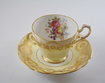 May include: A yellow teacup and saucer with a gold trim and floral design. The cup has a white interior and a floral pattern with a pear and other fruits. The saucer has a gold trim and a floral pattern.
