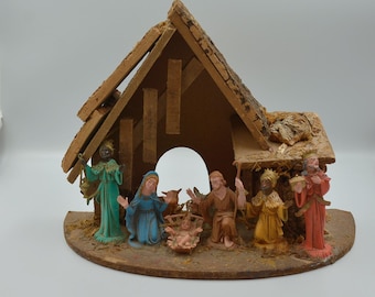 Op de afbeelding: Een kerststalset met een houten stalstructuur met een dak van boomschors. De figuren omvatten een centraal kind in een kribbe, Maria in het blauw, Jozef in het bruin en drie figuren in groene, gele en rode gewaden die ernaast staan.