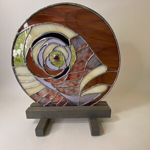 May include: A stained glass art piece with a swirling pattern of brown, white, green, and purple glass. The piece is mounted on a wooden stand.
