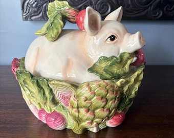 Op de afbeelding: Een keramische pot met deksel in de vorm van een varken dat rust in een kom versierd met gebeeldhouwde groenten. Het varken is lichtroze met een radijs op zijn rug. De basis bevat groene artisjokken en rode radijzen met gedetailleerde bladeren.