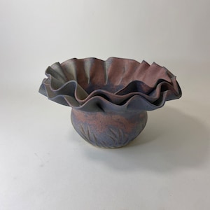 May include: A ceramic vase with a ruffled rim and a purple and pink glaze. The vase has a textured surface with a design of vertical lines.
