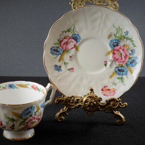 May include: A white teacup and saucer with a floral pattern featuring pink, blue, and yellow flowers. The saucer has a scalloped edge and the teacup has a gold rim.