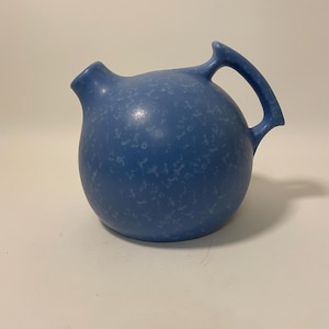 May include: A blue ceramic jug with a speckled glaze and a rounded body. The jug has a single handle and a spout.