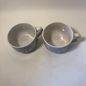 Puede incluir: Dos tazas de sopa de cer&aacute;mica blanca con asas. Las tazas tienen forma redondeada y un interior de color claro. El exterior de cada taza presenta un patr&oacute;n repetido de texto manuscrito y peque&ntilde;as ilustraciones en color azul oscuro.
